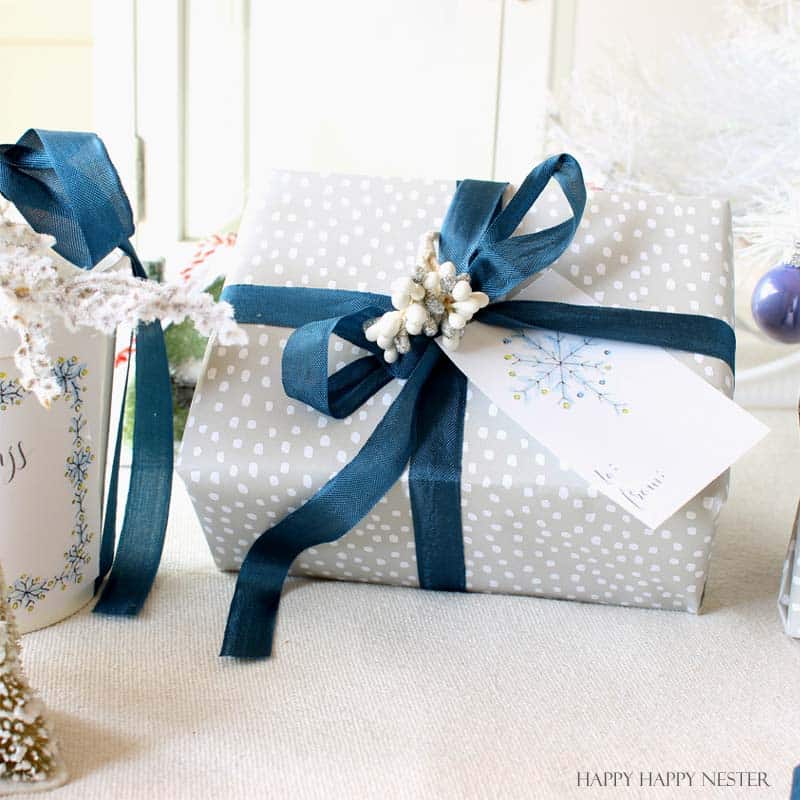 A beautifully wrapped gift with gray and white polka dot paper, adorned with a dark blue ribbon and a small bouquet of white berries. A cute Christmas tag featuring snowflake designs is attached, completing the festive, wintry setting with white decorations in the background.