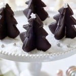 A white cake stand holds several chocolate Christmas tree-shaped cakes, resembling delightful Christmas tree cookies. These treats are adorned with small white stars on top, surrounded by a dusting of powdered sugar and silver sprinkles. Festive decorations blur beautifully in the background.