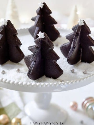 A white cake stand holds several chocolate Christmas tree-shaped cakes, resembling delightful Christmas tree cookies. These treats are adorned with small white stars on top, surrounded by a dusting of powdered sugar and silver sprinkles. Festive decorations blur beautifully in the background.