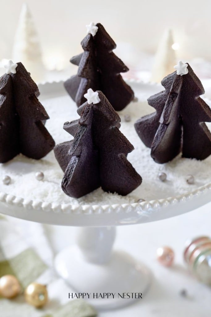 A white cake stand holds several chocolate Christmas tree-shaped cakes, resembling delightful Christmas tree cookies. These treats are adorned with small white stars on top, surrounded by a dusting of powdered sugar and silver sprinkles. Festive decorations blur beautifully in the background.