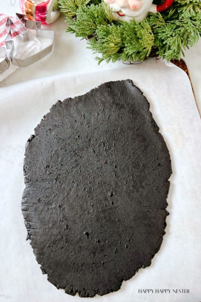 A sheet of black cookie dough is rolled out on parchment paper. In the background, there's a festive wreath with a Santa Claus decoration and some Christmas tree cookie cutters.