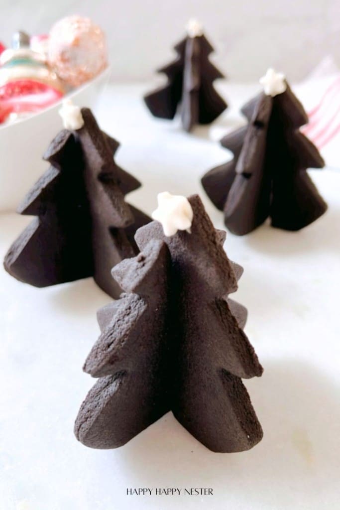 Four chocolate Christmas tree-shaped treats are arranged on a white surface. Each tree is topped with a small white star. In the background, there is a dish containing festive ornaments.