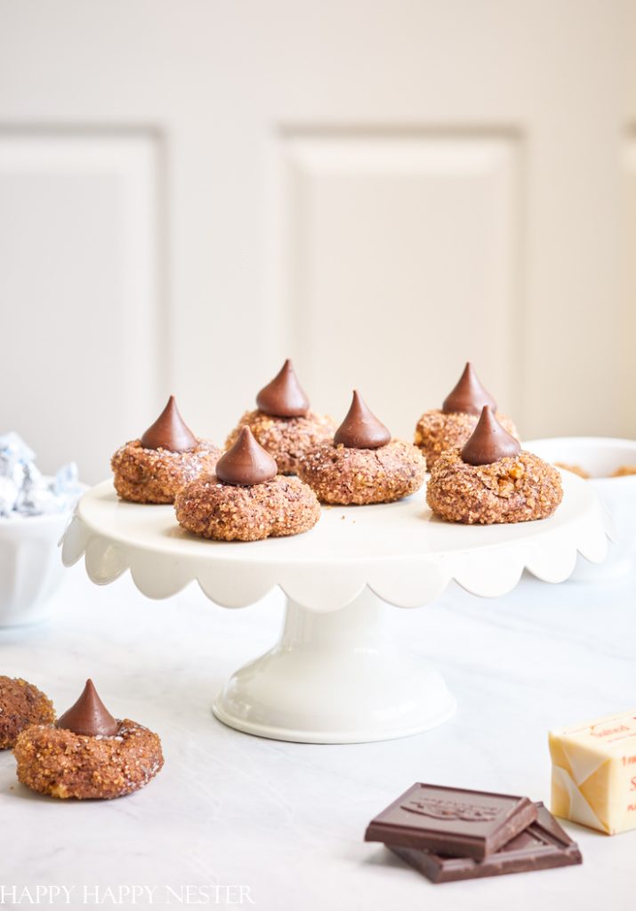 Cookies topped with chocolate kisses are arranged on a white cake stand, making them perfect for Christmas treat recipes. The background is softly blurred, featuring light tones and a piece of chocolate in the foreground.