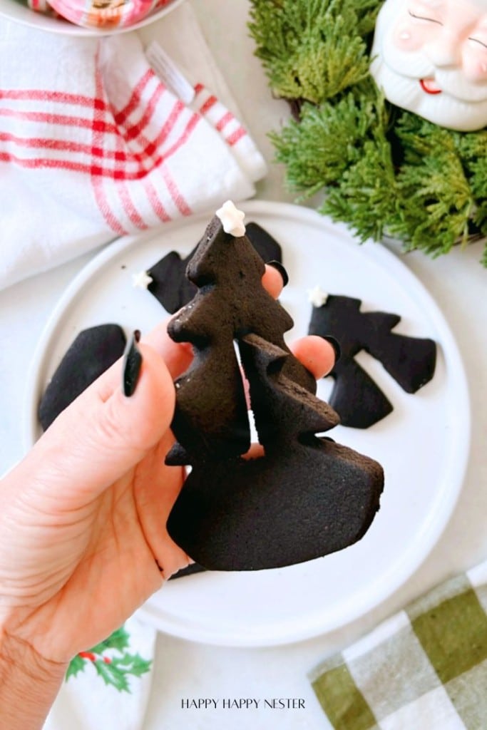 A person holds a black tree-shaped cookie with a white star on top. In the background, a white plate with similar cookies, a Santa-themed container, a green wreath, and a red-striped towel are visible.