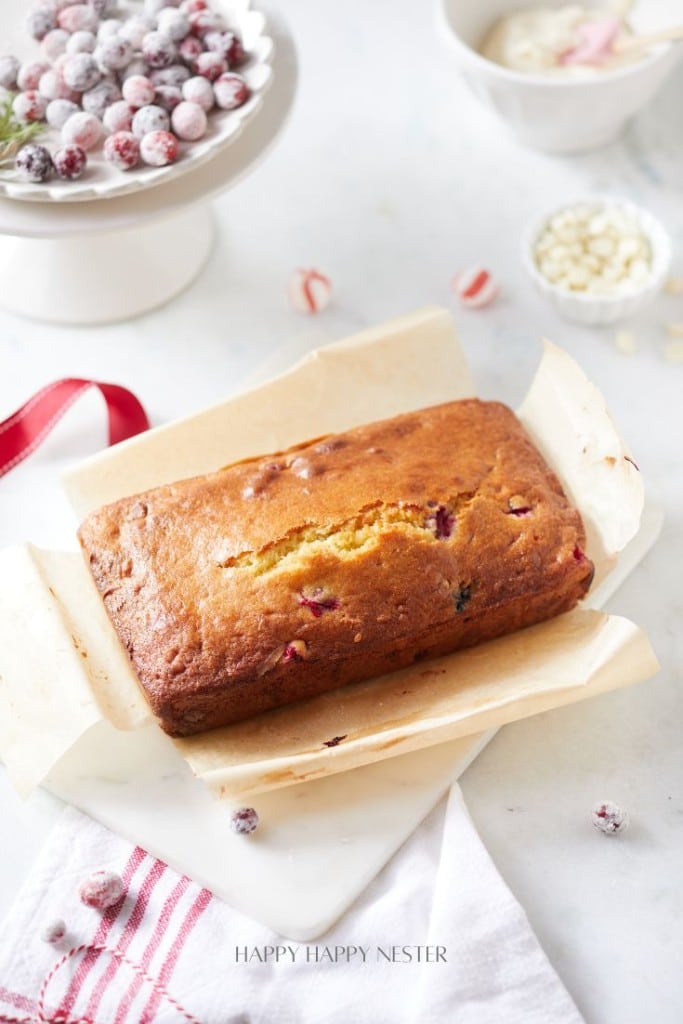 A loaf of cranberry bread sits on parchment paper. Surrounding it are sugared cranberries on a stand, a bowl with a creamy mixture, and a white dish with white chocolate chips. A red-striped cloth napkin is nearby.