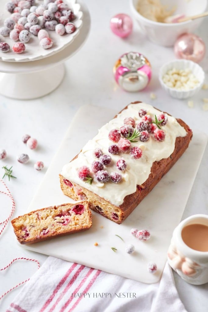 A loaf cake topped with white frosting and sugared cranberries on a marble board. A slice is cut from the cake. Surrounded by loose cranberries, a red-striped cloth, and festive ornaments on a white surface.