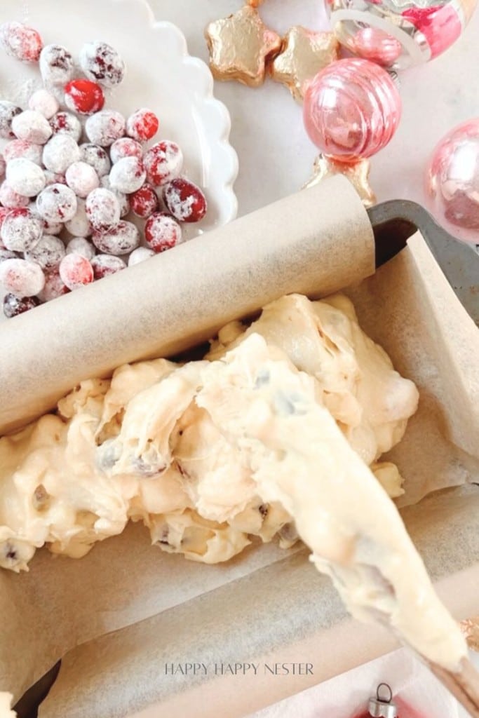 Baking scene with cookie dough in a parchment-lined tray, a plate of sugared cranberries, and festive decorations like pink ornaments and star-shaped cookies in the background.