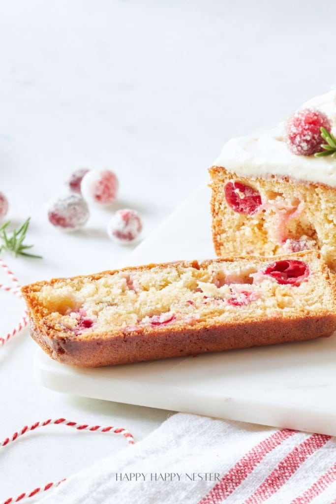 A close-up of a loaf cake with a slice cut, showcasing cranberries inside. The cake is topped with frosting and sugared cranberries. A few cranberries and a sprig of rosemary lie beside the cake on a white surface.