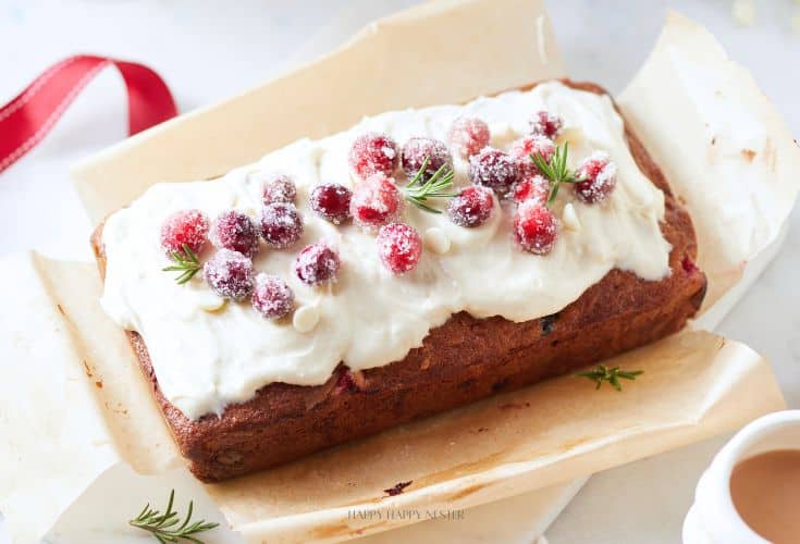 A rectangular holiday cake topped with white frosting, garnished with sugared cranberries and small rosemary sprigs. The cake is placed on parchment paper, and there's a red ribbon in the background.