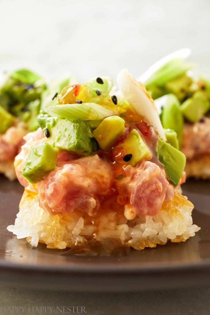 Close-up of a sushi dish with a crispy rice base, perfect for new year's recipes. It features diced avocado, green onions, black sesame seeds, and a drizzle of sauce over fresh raw fish. The vibrant colors highlight the fresh ingredients.