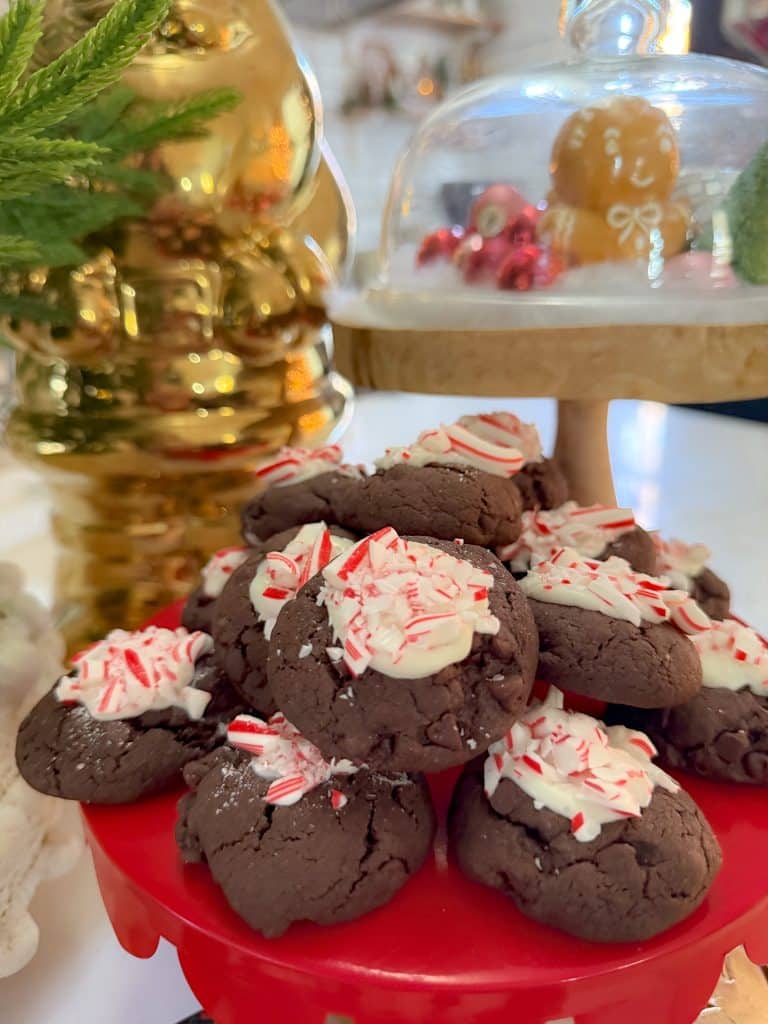 A festive display of chocolate peppermint cookies topped with crushed candy canes on a red plate captures the holiday spirit. In the background, a glass cake stand holds a gingerbread house, flanked by Christmas tree cookies and a gleaming gold holiday decoration.