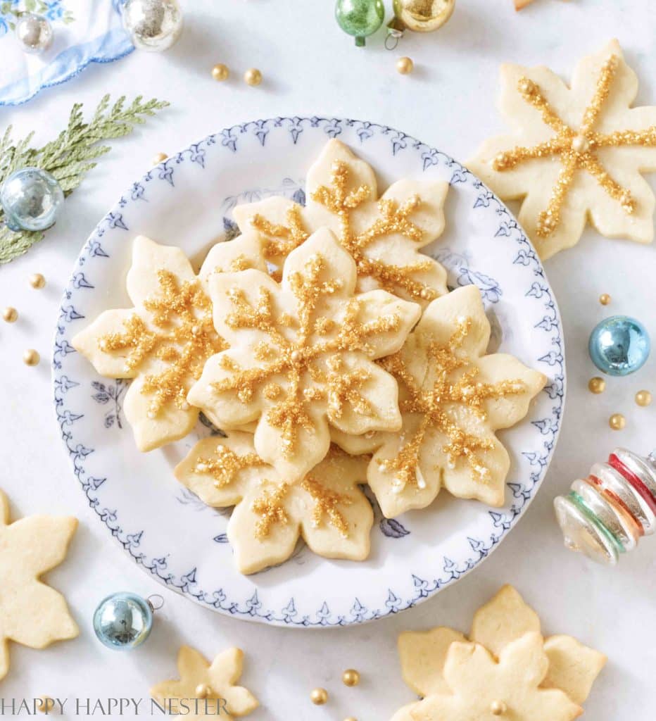 A plate of snowflake-shaped cookies, made from the finest Christmas treat recipes, is beautifully decorated with gold glitter sprinkles. Surrounding the plate are small Christmas ornaments and more cookies, all elegantly arranged on a light-colored surface.