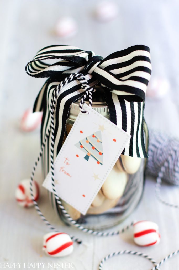 A jar filled with cookies is adorned with a black and white striped ribbon and cute Christmas tags featuring a festive tree. Peppermint candies are scattered around, along with a spool of twine, creating a delightful holiday-themed scene.