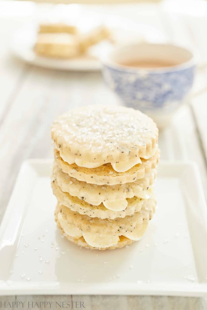 A stack of lemon poppy seed cookies with icing sits on a white square plate, resembling a delightful entry in Christmas treat recipes. A cup of tea in a patterned cup and more cookies are blurred in the background.