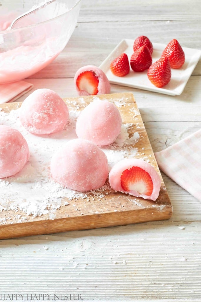 Pink mochi balls filled with strawberries are artfully arranged on a wooden board dusted with powdered sugar, making for delightful New Year's recipes. A few whole strawberries rest on a small white plate in the background, and a cut mochi reveals the fresh strawberry within.