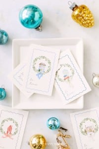 Christmas gift tags with wreath designs and pastel house illustrations on a white plate. Surrounding the plate are ornaments in blue, gold, and silver, including a pinecone and ball shapes, on a light surface.