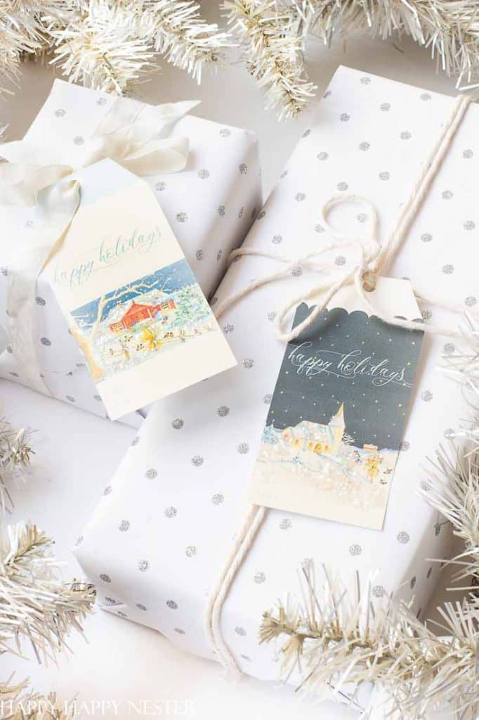 Two white gift boxes wrapped in silver polka dot paper are decorated with cute Christmas tags. One tag features a snow-covered tree and house, while the other shows a snowy church scene. Both read "Happy Holidays," with silver garland beautifully surrounding the gifts.