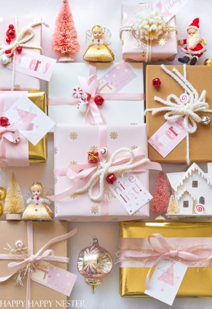 A collection of beautifully wrapped gifts with pink and gold ribbons, topped with decorative ornaments like angels, a Santa figure, and miniature trees. Some presents have cute Christmas tags adorned with festive messages.