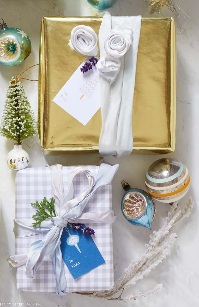 Two beautifully wrapped gifts on a marble surface. The top gift, adorned with cute Christmas tags, is in gold paper with a white ribbon. The bottom one features checkered paper with a blue tag and plant decoration. Nearby are ornaments and a small Christmas tree, enhancing the festive charm.