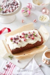 A festive cranberry bread recipe loaf is topped with white frosting and sugared cranberries, garnished with rosemary sprigs. It rests on parchment paper, surrounded by holiday decorations like ornaments and a bowl of cranberries. A cup of tea waits invitingly on the side.