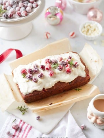 A festive cranberry bread recipe loaf is topped with white frosting and sugared cranberries, garnished with rosemary sprigs. It rests on parchment paper, surrounded by holiday decorations like ornaments and a bowl of cranberries. A cup of tea waits invitingly on the side.