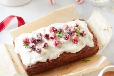 A festive cranberry bread recipe loaf is topped with white frosting and sugared cranberries, garnished with rosemary sprigs. It rests on parchment paper, surrounded by holiday decorations like ornaments and a bowl of cranberries. A cup of tea waits invitingly on the side.