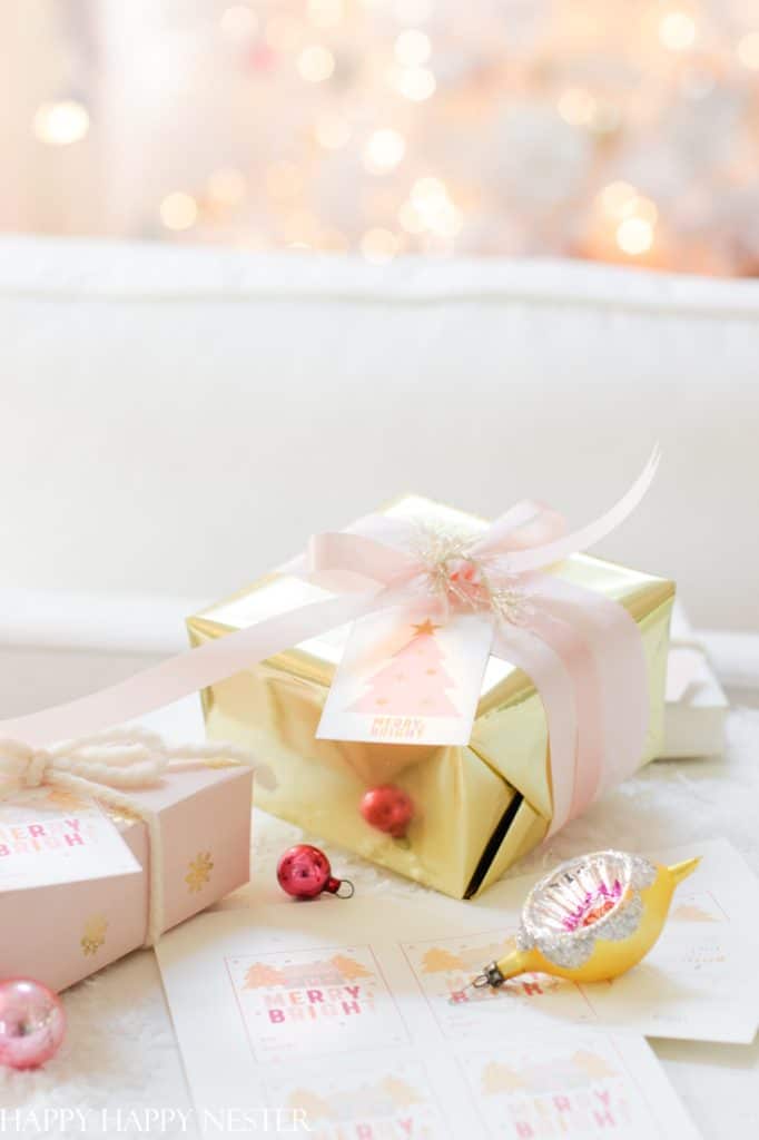 A festive scene with Christmas presents wrapped in gold and white paper, adorned with pink ribbons. Cute Christmas tags reading "Merry & Bright" are attached to the gold box. Small ornaments and a glowing tree twinkle softly in the background.