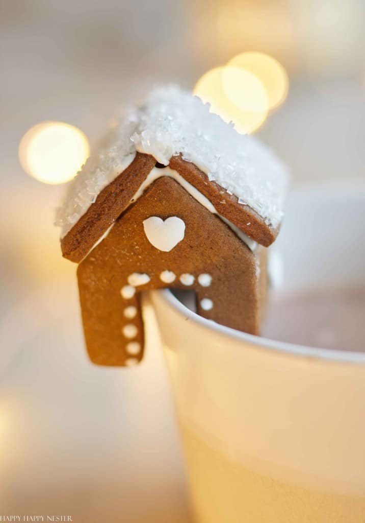 A charming gingerbread house with white icing and a heart detail perches on the rim of a cup. The roof, dusted with sugar, evokes the coziness of Christmas treat recipes, while soft, warm lights glow in the background.