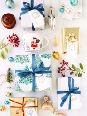 An overhead view of a festive arrangement featuring wrapped gifts with blue and gold ribbons, printable holiday tags, a Santa-themed mug, decorative ornaments, holly berries, and small holiday figurines on a light background.