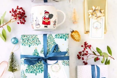 An overhead view of a festive arrangement featuring wrapped gifts with blue and gold ribbons, printable holiday tags, a Santa-themed mug, decorative ornaments, holly berries, and small holiday figurines on a light background.