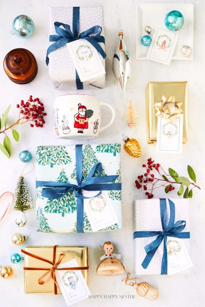 An overhead view of a festive arrangement featuring wrapped gifts with blue and gold ribbons, printable holiday tags, a Santa-themed mug, decorative ornaments, holly berries, and small holiday figurines on a light background.