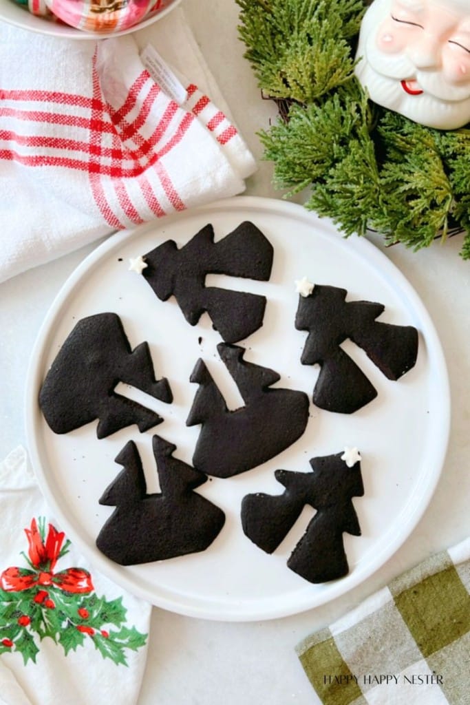 A plate with Christmas tree cookies, their festive black shapes adding charm to the white surface, is surrounded by holiday-themed napkins, greenery, and a cheerful Santa Claus mug.