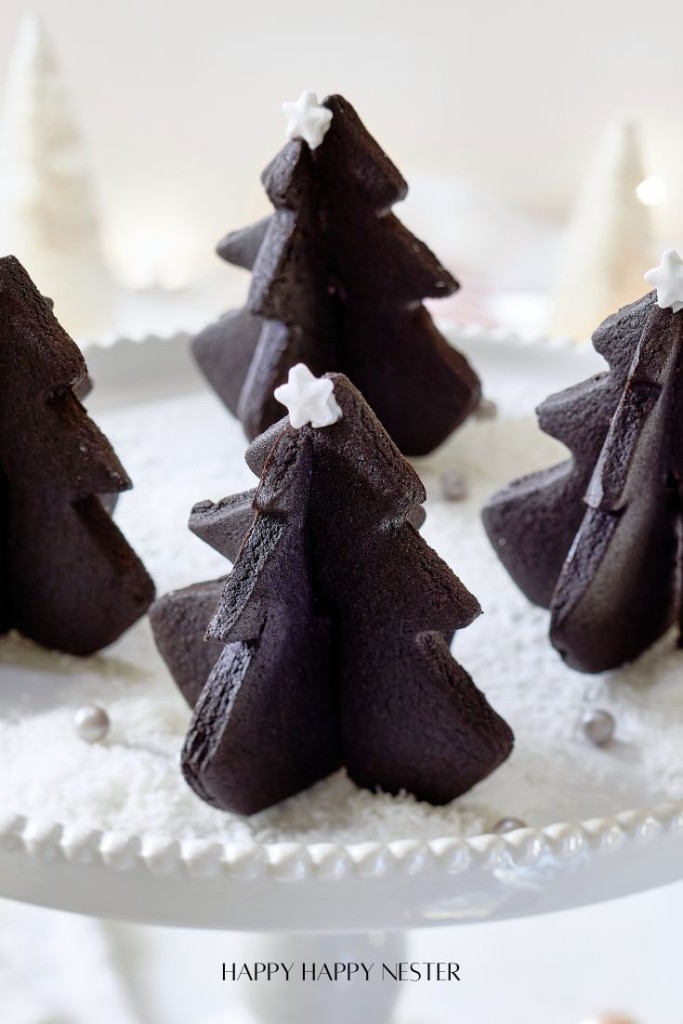 Chocolate Christmas tree-shaped cakes with star decorations on top are displayed on a white cake stand, surrounded by fake snow and small silver balls. The background is softly blurred.