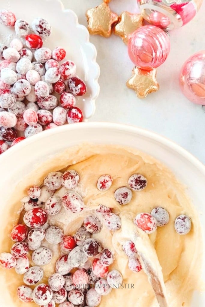 A bowl of cake batter with sugared cranberries being mixed in, inspired by a delightful cranberry bread recipe. Nearby, a dish of extra sugared cranberries and pink ornaments shaped like stars and spheres are displayed on a light surface.