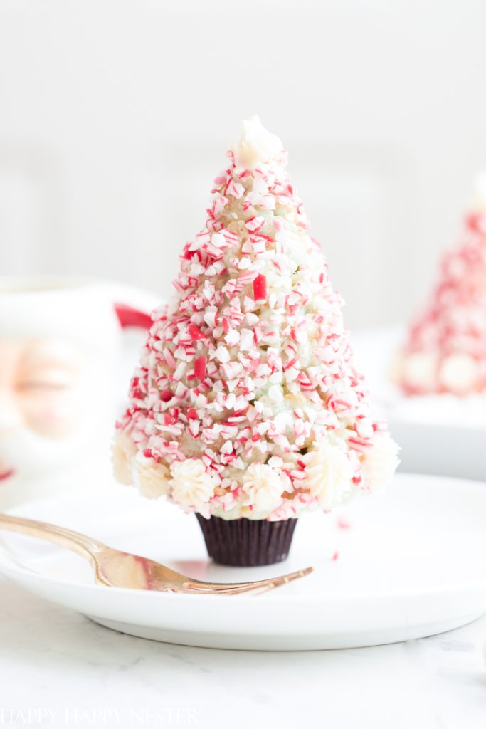 A delightful cupcake, styled like a Christmas tree and adorned with white and red crushed candy canes, sits on a white plate. A gold fork rests beside it. The blurred background suggests a collection of similar festive treats, all inspired by the best Christmas treat recipes.