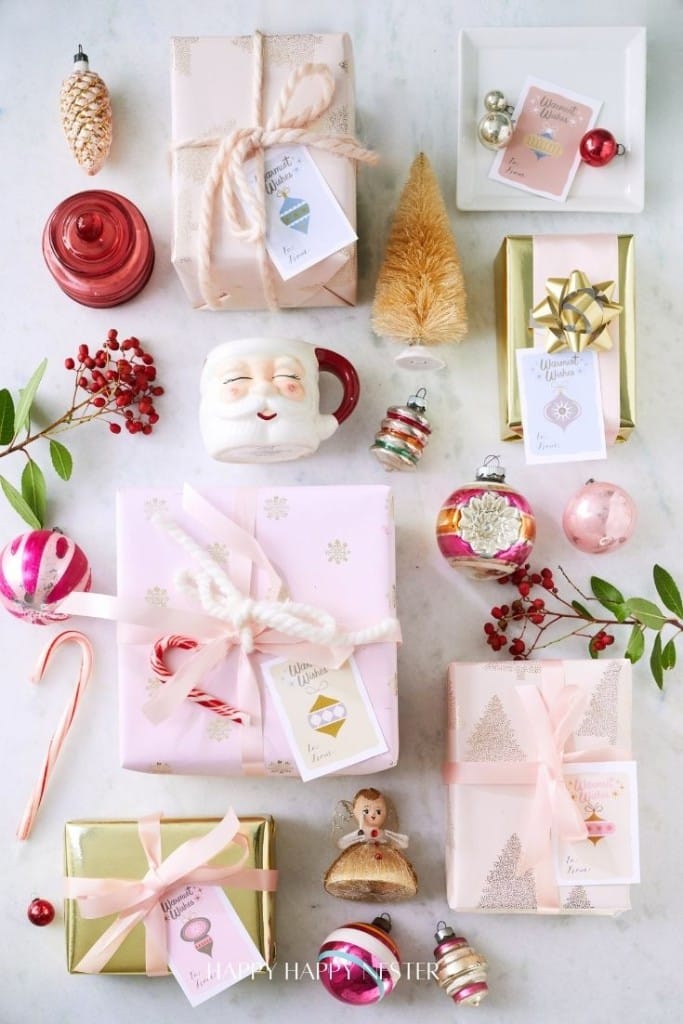 A festive arrangement features gift boxes wrapped in pink and gold paper with ribbons, surrounded by ornaments, a Santa mug, a small gold tree, and red berries. Cute Christmas tags display cheerful holiday messages on the presents.