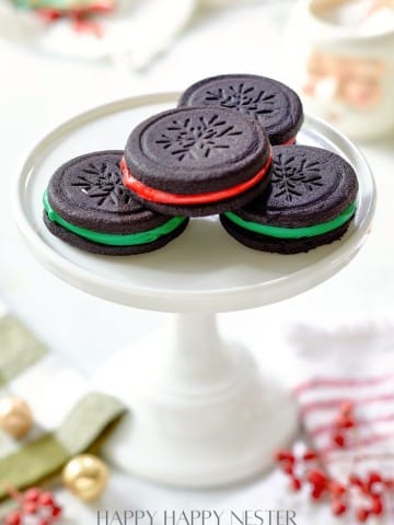 A white cake stand showcases four Oreo cookies with intricate snowflake patterns. Two boast rich red filling, while the others reveal a vibrant green. The backdrop brims with festive holiday decor, including ribbons and sparkling ornaments, setting the perfect scene for an Oreo cookie recipe delight.