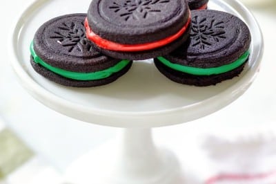 A white cake stand showcases four Oreo cookies with intricate snowflake patterns. Two boast rich red filling, while the others reveal a vibrant green. The backdrop brims with festive holiday decor, including ribbons and sparkling ornaments, setting the perfect scene for an Oreo cookie recipe delight.