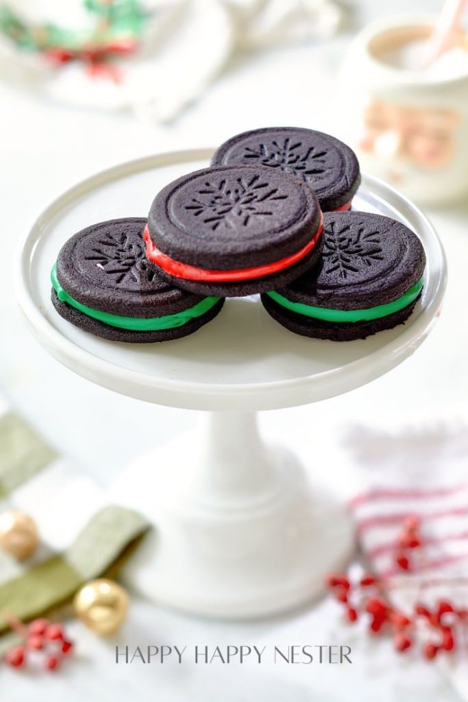 Four chocolate sandwich cookies with festive red and green fillings are stacked on a white cake stand, exemplifying perfect Christmas treat recipes. A mug with a Santa design and holiday decorations are blurred in the background.