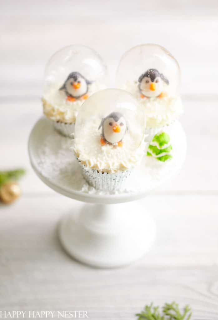 Three cupcakes topped with miniature penguin figures inside clear gelatin domes are displayed on a white cake stand. Resembling snow, the white frosting captures the holiday spirit, making these delightful treats a charming addition to your Christmas treat recipes collection, complete with a small green decoration.