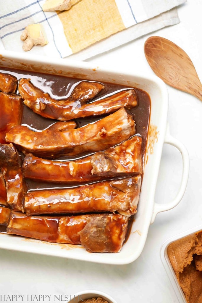 A white baking dish filled with uncooked ribs marinated in a dark brown sauce sits ready for your New Year's recipes. The dish rests on a white surface, accompanied by a wooden spoon and cloth napkin, while small bowls with seasonings are visible nearby.