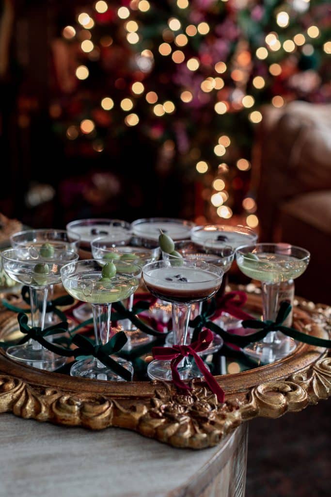 Champagne glasses with green olives arranged on an ornate tray, surrounded by red and green ribbons. A snowglobe glistens nearby, while blurred Christmas lights in the background create a festive atmosphere.