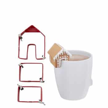 A white mug filled with a hot beverage features a small gingerbread house resting on its rim, evoking the charm of Christmas treat recipes. To the left, three red metal cookie cutters in house-like shapes are displayed in a row.