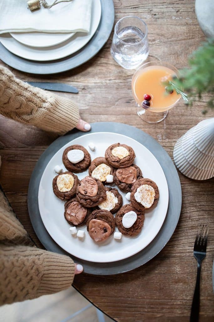 A person in a knitted sweater presents a plate of chocolate cookies adorned with marshmallows, biscuits, and festive Christmas tree cookies. The table is set with a glass of orange drink and a decorative vase, creating a cozy and inviting atmosphere.