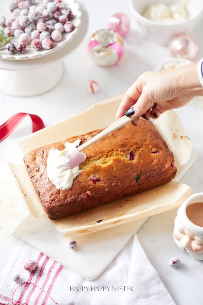 A person is crafting a delightful cranberry bread recipe, spreading white frosting on the loaf with a spatula. The cake rests on parchment paper surrounded by a cup of coffee, a red-striped cloth, sugared cranberries in a bowl, and decorative ornaments.