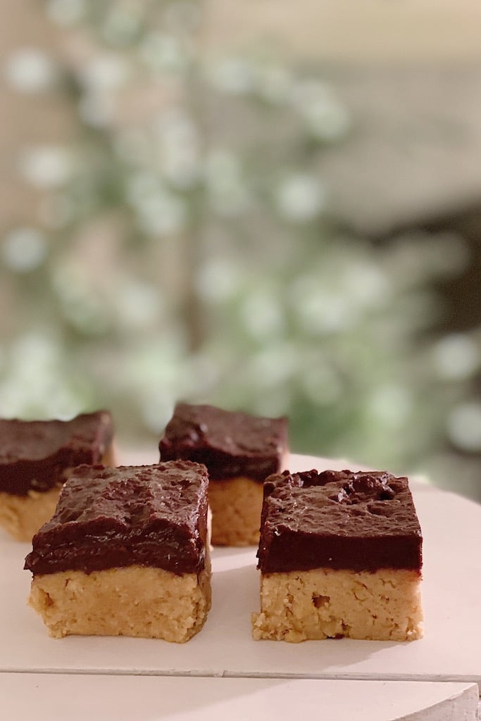 Close-up of four chocolate-topped peanut butter bars on a white surface, perfect for New Year's recipes. The background is softly blurred with muted green and beige tones, creating a warm, inviting atmosphere.