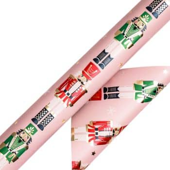 A roll of gift wrap featuring cute nutcracker soldiers in green and red uniforms on a pink background sprinkled with small stars. The paper is partially unrolled, perfect for pairing with cute Christmas tags to complete your festive presentation.