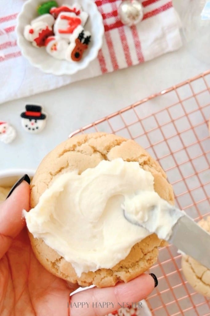 A hand holding a cookie spread with white frosting using a knife. In the background, there are festive decorations, including small bowls with holiday-themed treats like Santa and snowman designs, on a red and white-striped cloth.