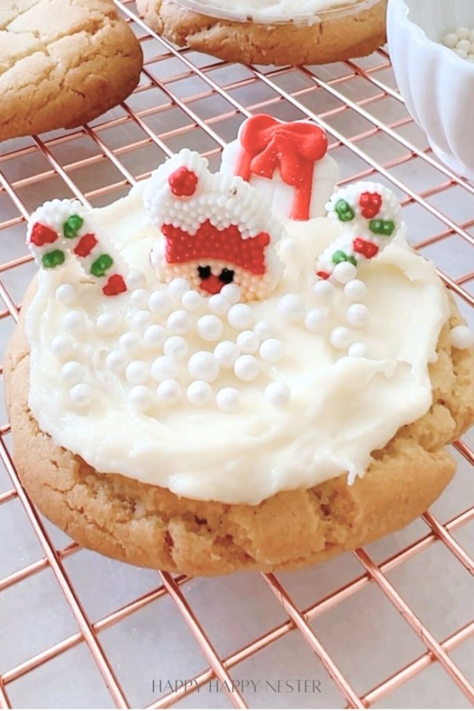 A festive cookie topped with white frosting, decorated with small white candy pearls, a snowman face, and tiny candy mittens and hat in red, green, and white colors, placed on a cooling rack.
