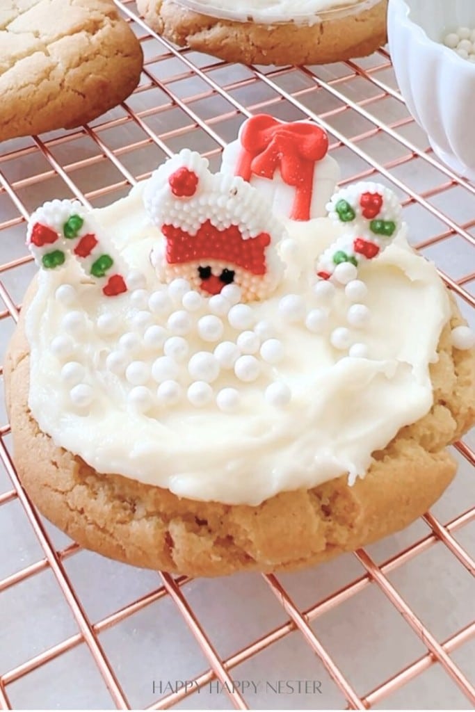 A Christmas-themed cookie with white frosting and small white sugar pearls. It features a Santa decoration, showing only his hat, gloves, and a small nose, as if emerging from the frosting. The cookie rests on a cooling rack.